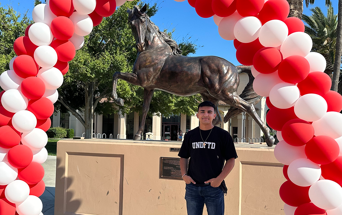 A young man in a black t-shirt and jeans stands under a balloon arch and the Bronco statue.