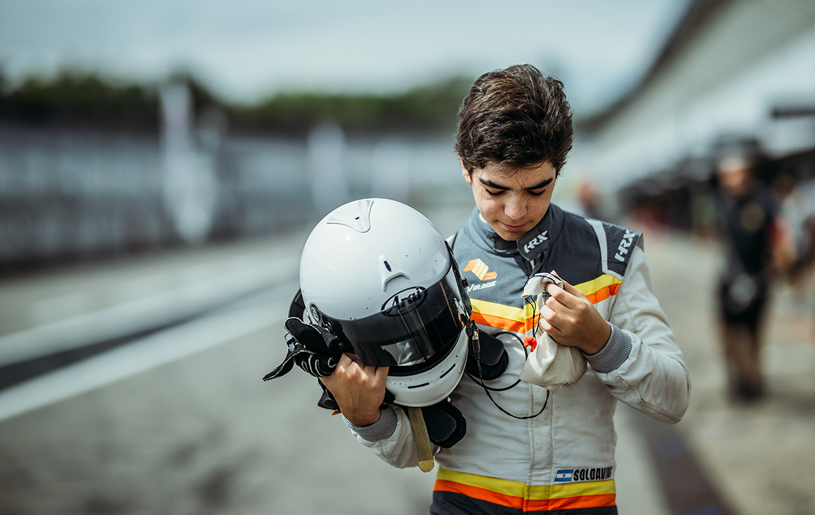 A young man in a racecar driver suit holding a racing helmet walks on a racecar track.