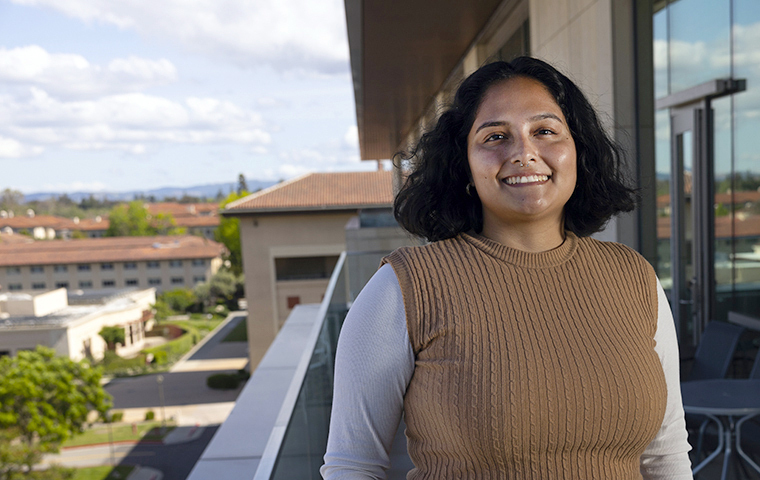 A student stands on a balcony with the sky and campus behind her.