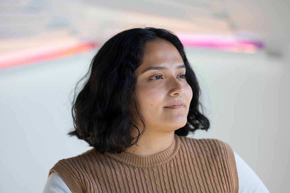 A closeup of a student standing in front of a pink neon light on a white wall.