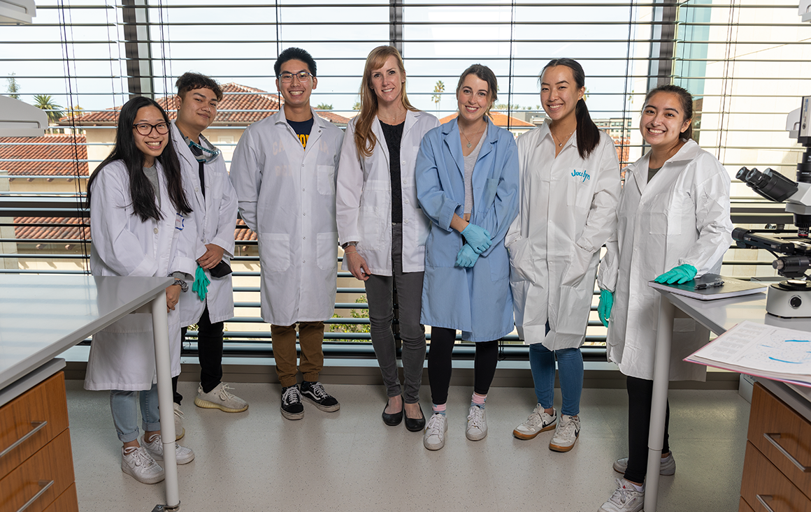 A group of students and a professor wearing lab coats stand shoulder to shoulder in a lab.