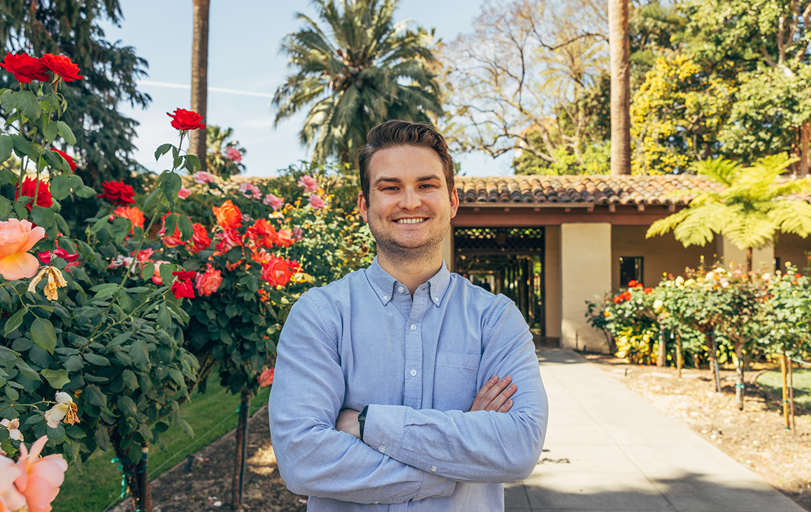 A man in a blue button-up, crossing his arms across his chest in front of a lush rose garden, with palm trees and an adobe entryway in the background.