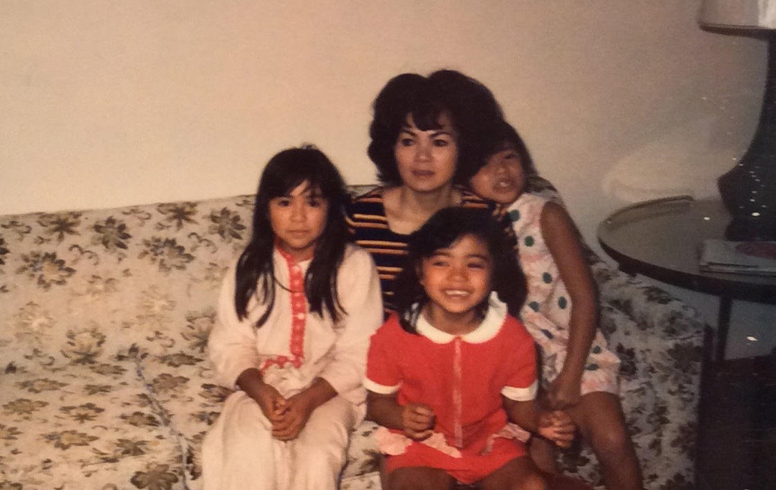 A japanese woman sits on a white couch with three daughters sitting on her lap.