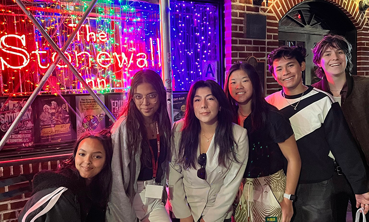 A group of students stand outside of a neon-lit sign for the Stonewall Inn.