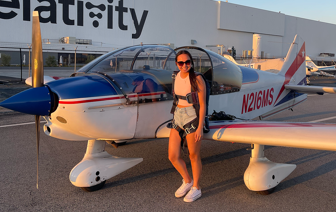 A young woman in a safety harness stands casually beside a small propellor plane.