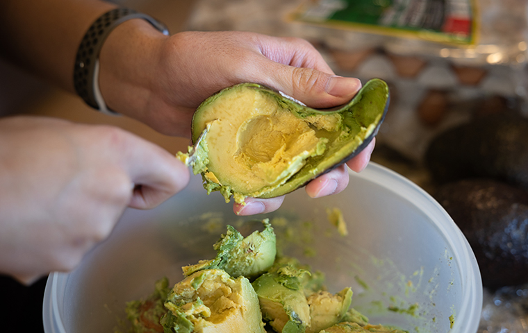 Hands with knife preparing an avocado over a bowl full of avocado