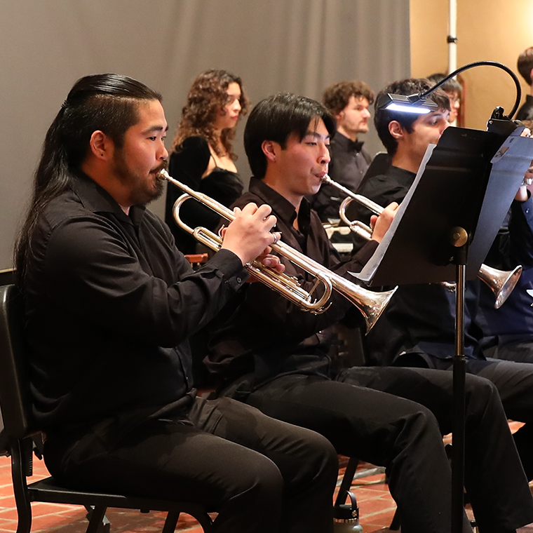 A group of musicians wearing black performing in a concert, Jared Shimada at the front playing a trumpet.