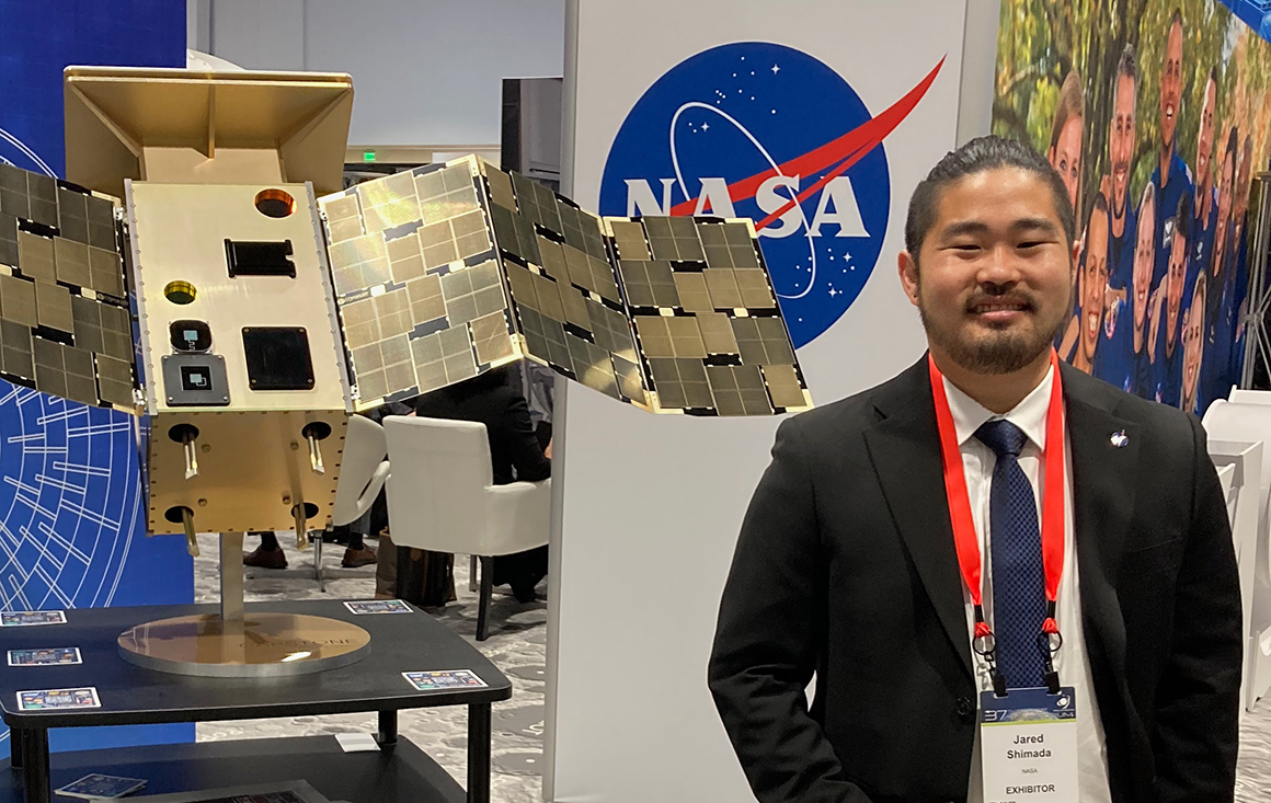 An Asian man wearing a suit, Jared Shimada, stands beside a gold model of a space probe in a conference space.