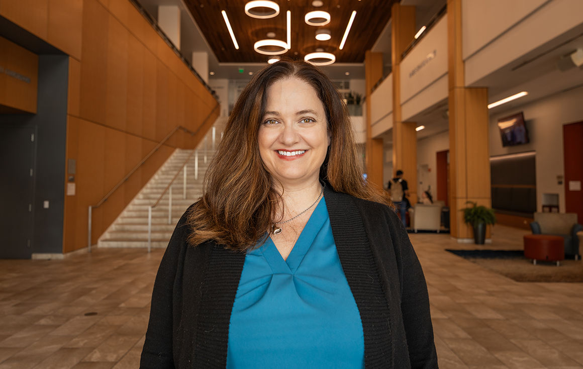 A woman in a blue blouse and black blazer stands in the atrium of the Santa Clara School of Law, circular ceiling lights shine dramatically in the background.