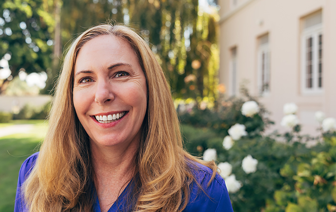 A blonde woman in a blue blouse smiles warmly, standing in a rose-filled garden by a willow tree.