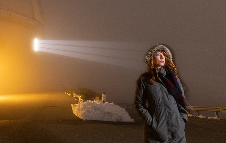 A woman standing outside an observatory at night, with a beam of light shining through the dark at the woman's head.