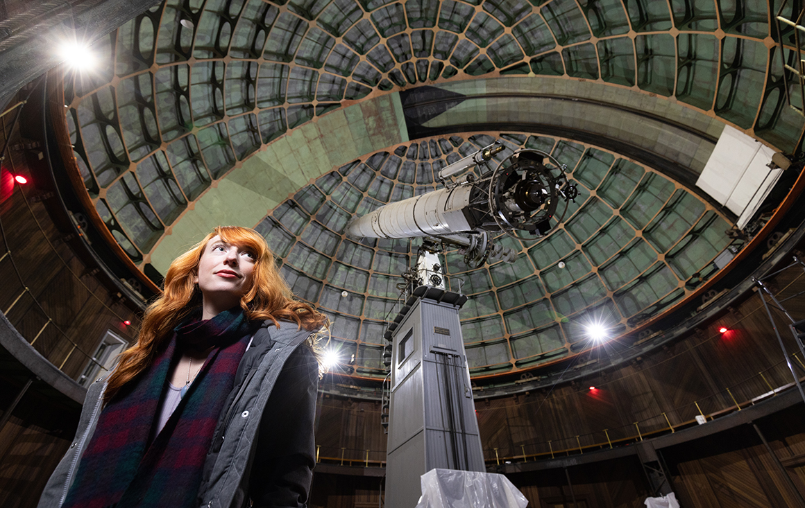 A woman standing below a large astronomical telescope.