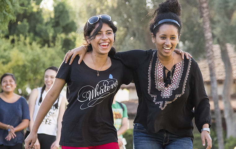 Two women students walk across campus with their arms over their shoulders