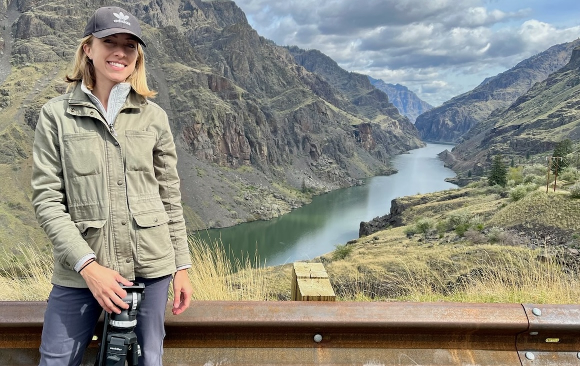 A woman, Maggie Beidelman, dressed in a cap and jacket stands with a tripod in front of Hells Canyon gorge.