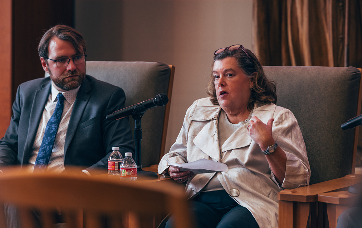 A woman in an armchair speaks into a microphone, and a man listening in a seat beside her.