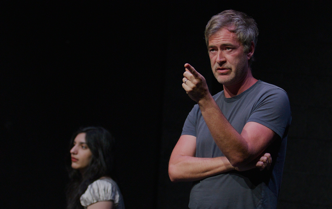 A man in a black box theater points off-camera with a student standing behind him.