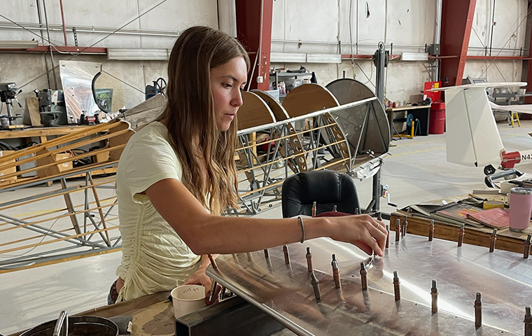 A young woman, Milaina Almonte, screws in metal pegs on a wing-like structure, with a bi-plane frame in the background.