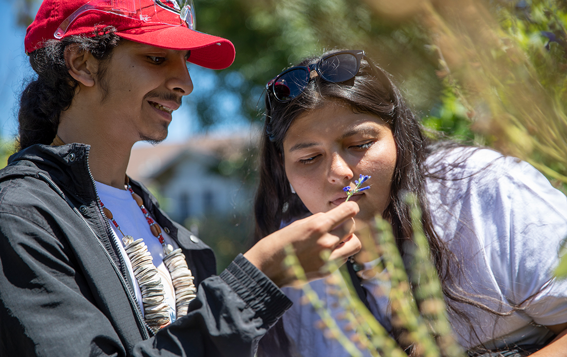 Two young people—a boy and a girl—lean in to look at a plant in a garden.