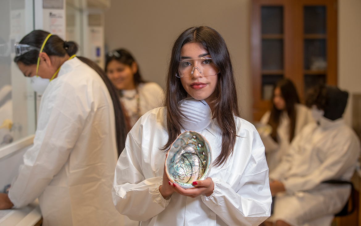 A young woman in a lab coat and protective goggles holds an abalone shell. Behind her, other young people in similar clothes are working in a lab.