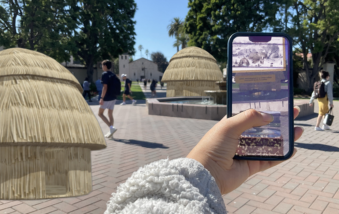 A hand holds a phone up to the Santa Clara University campus, with digitally rendered tule houses made by reeds overlaid onto the campus landscape.