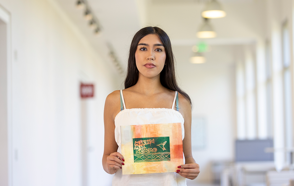 A young woman, Isabella Gomez, holds up a dark green linocut print of a hummingbird on pink and yellow-stained paper.