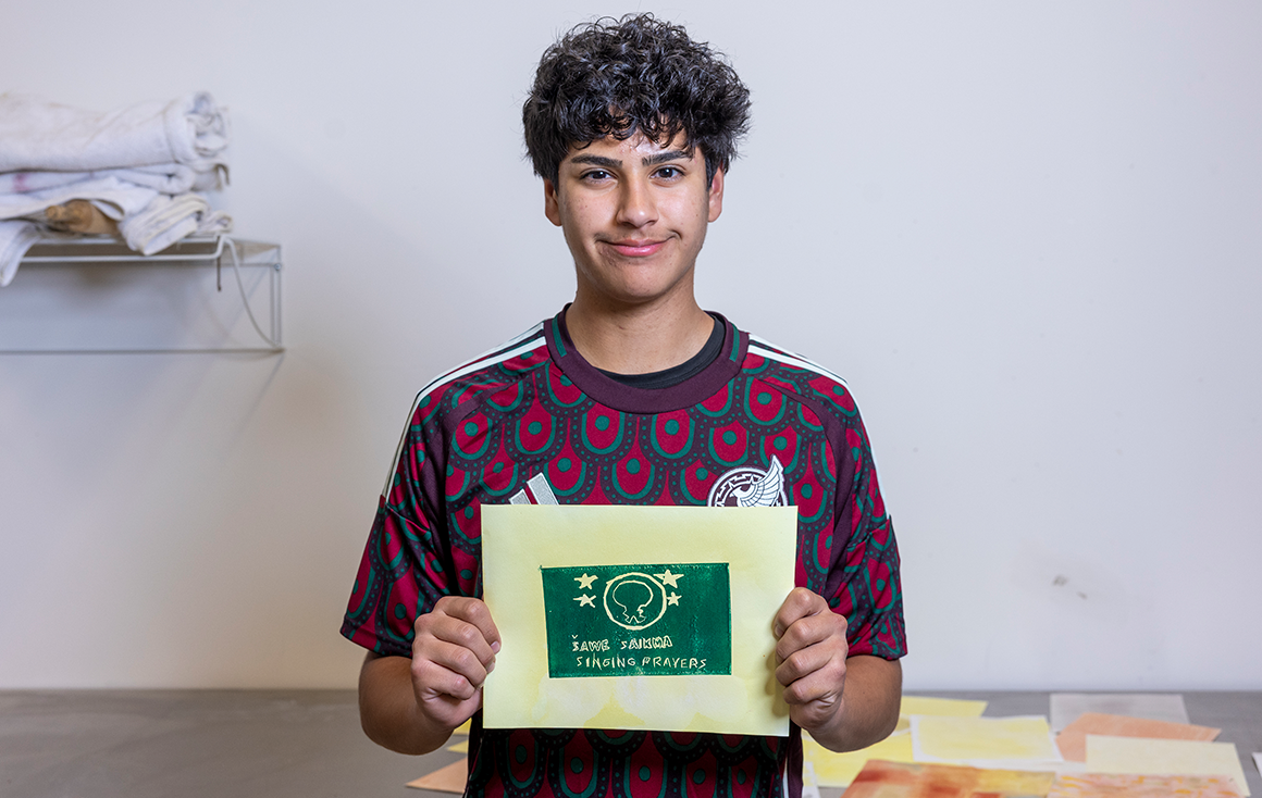 A young man, Lucas Arellano, holds up a dark green print with an abstract figure and four stars on green-stained paper.