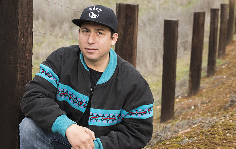 Author Tommy Orange kneeling in front of a rural fence (Photo by Elena Seibert) image link to story