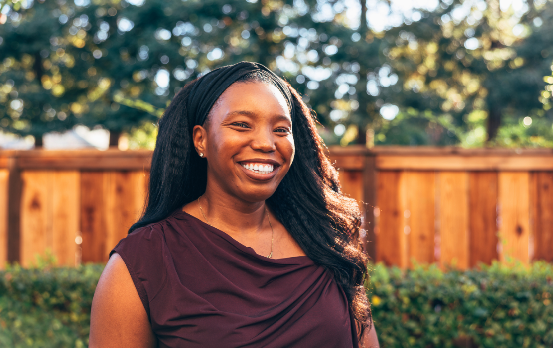 A black woman in a brown blouse stands in front of a wooden fence