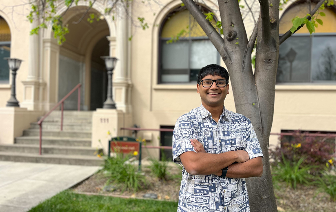 A young man in a patterned shirt stands arms crossed by a tree in front of an academic building.