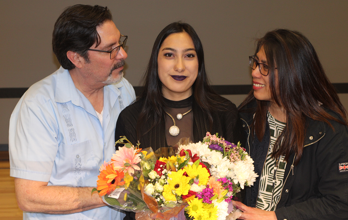 A young woman holds a bouquet of flowers, flanked by her adoring parents on either side.