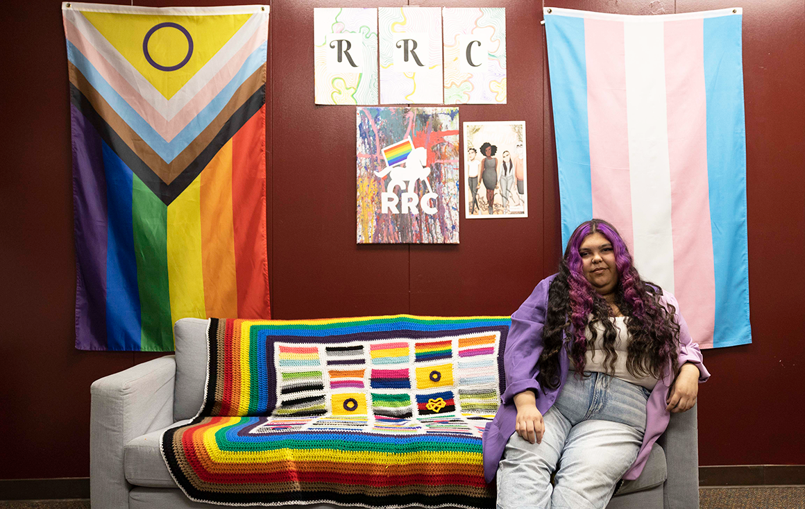 A student sits on a couch covered with a crocheted pride flag blanket, with two more LGBTQ+ pride flags behind her on the wall.