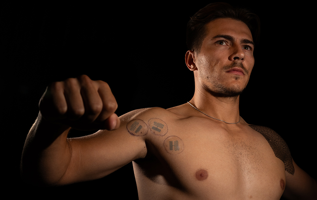 Nick Cmager with Microfluidic sensors adhered to his bare shoulder in a dark room with spotlight.