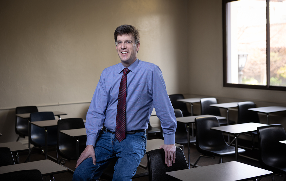 A professor sits on the edge of a student desk in a classroom surrounded by rows of other empty desks.