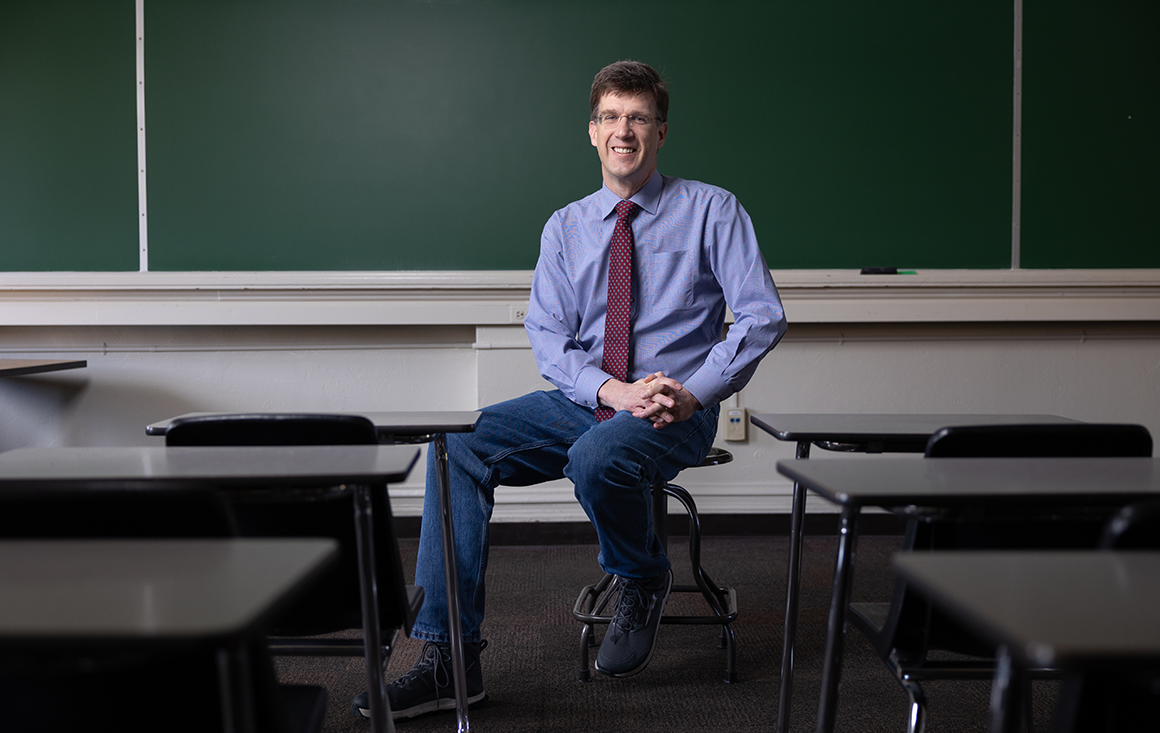 A professor sitting on a stool in front of a classroom chalkboard with empty student desks in the foreground.