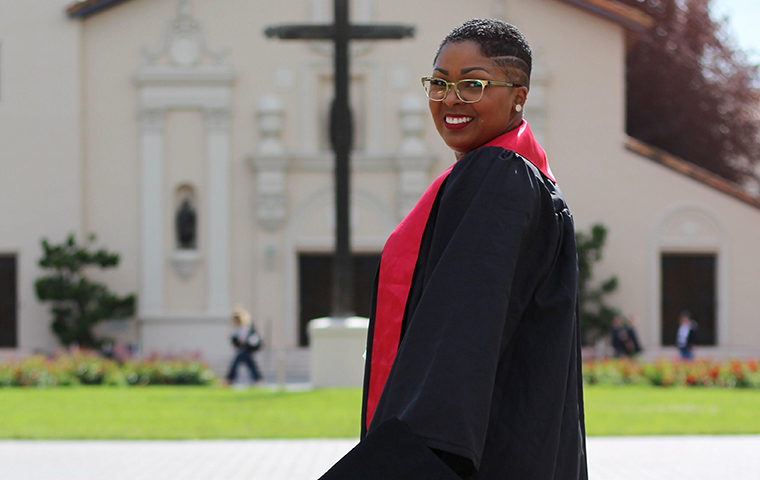 Tanesha looks at the camera over her shoulder with the Mission in the background. She wears her undergraduate gown and red ribbon.