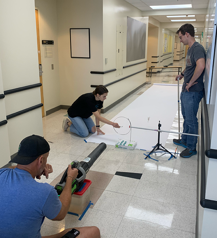 A professor aims a leafblower down a school hallway covered in white butcher paper. Two students stand ready to release seeds and measure distance.