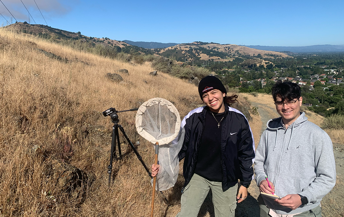 Two students in the grasslands of the Coyote Hills, holding a butterfly net and a notebook, with a film camera focused on a succulent on a rocky outcrop.