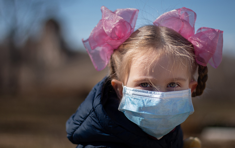 A young girl with two bows in her hair wearing a medical mask.