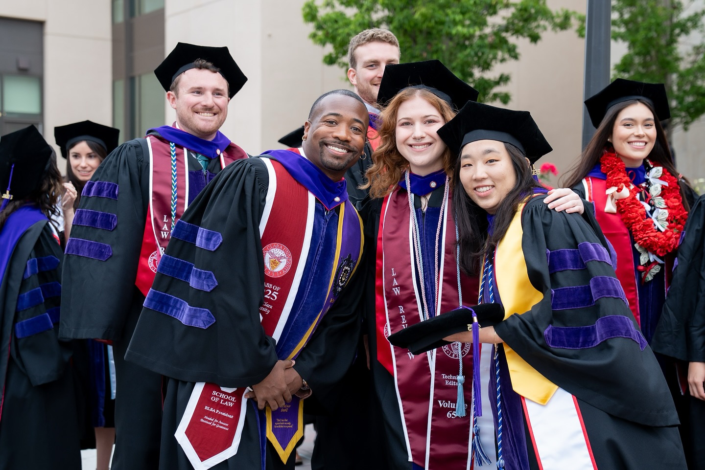 Five Santa Clara Law graduates in gowns and sashes posing for a photo