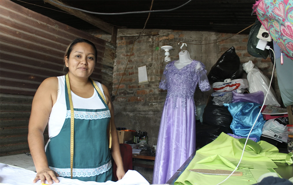 A woman wearing an apron and measuring tape stands in front of a table with garments on it