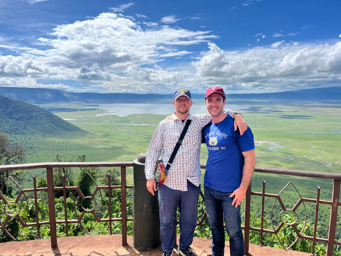 Two men smiling. Behind them is a large expanse of green grass and green plateaus with a body of water in the middle