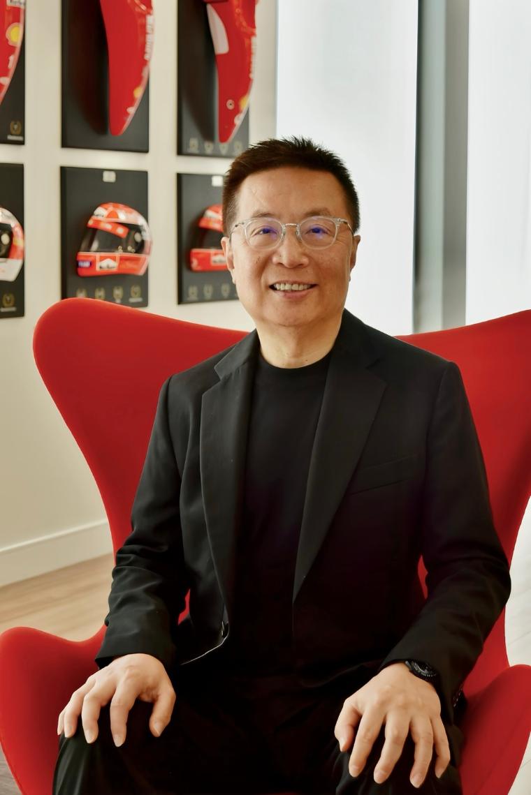 A man sits on a red chair with racecar helmets behind him. He is wearing a black suit and glasses.