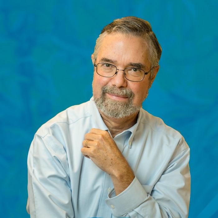 A man poses in front of a blue background. He wears glasses and has facial hair. One arm is across his body.