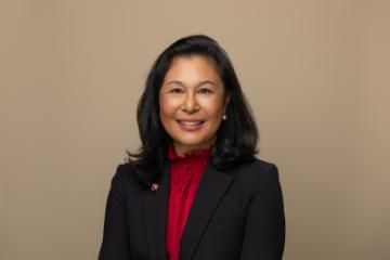 A woman wearing a red blouse and a black blazer smiles in front of a tan background.