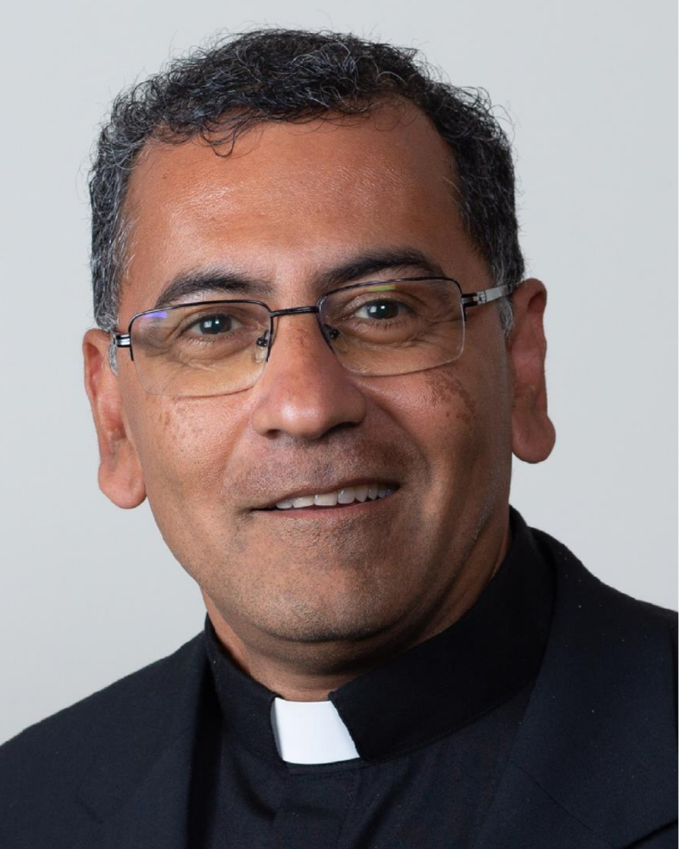 A man smiles in front of a light grey background. He wears a clerical collar and glasses.