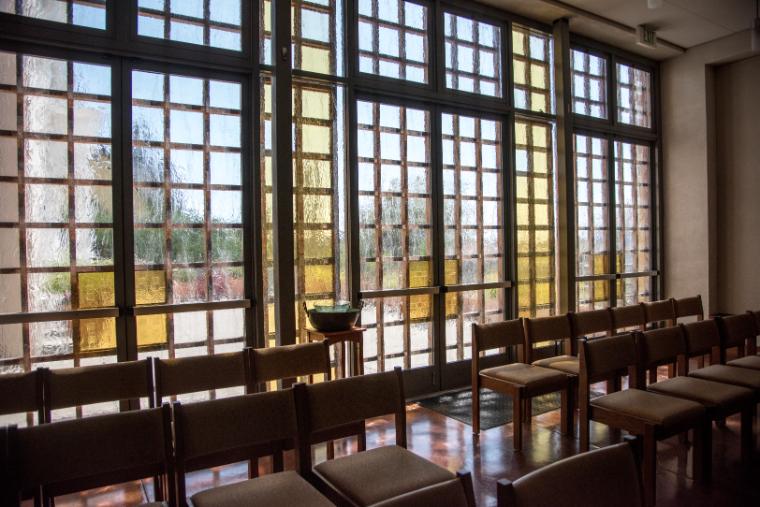 Empty chapel with wooden pews and large stained-glass windows.