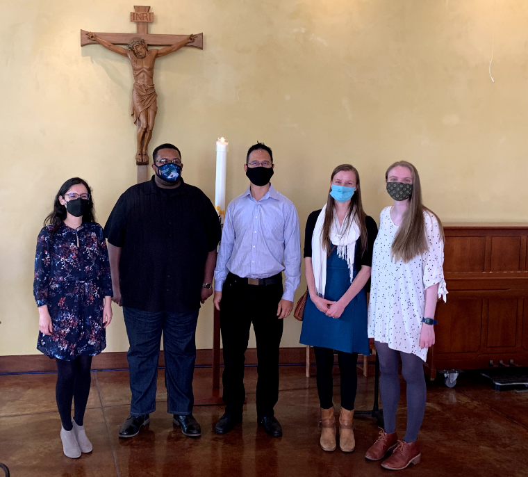 Five people wearing masks, standing in a church under a cross.