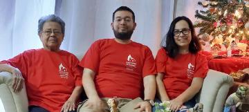 Three people in red shirts sitting on a couch near a Christmas tree.