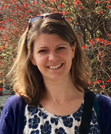 A woman smiling in front of a bush with red flowers.