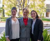 Three people posing outdoors with greenery and buildings in the background.